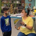 Colorful classroom scene with teacher and student wearing masks, engaging in a reading activity at Unicorn Jazz educational center. Bright, welcoming environment for learning and creativity.